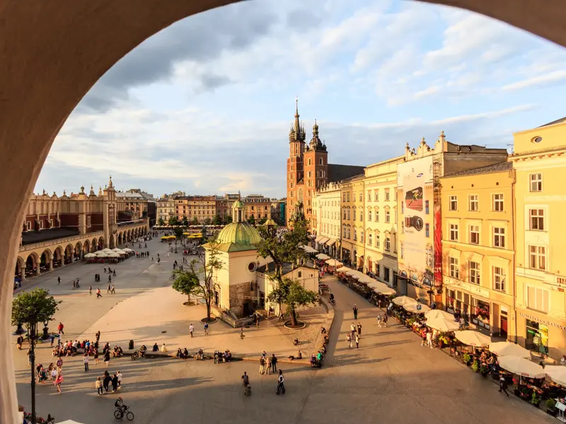 Blick durch einen Bogen auf den belebten Hauptring in Krakau mit den Tuchhallen und der Marienkirche in der Abendsonne.