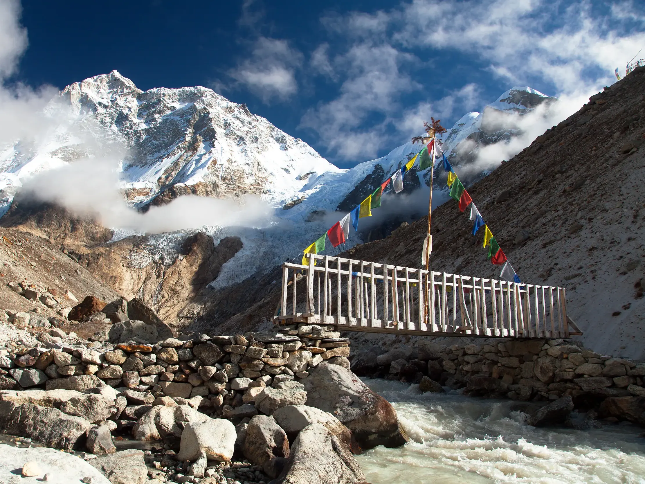 Eine Holzbrücke mit bunten Gebetsfahnen führt über einen Fluss beim Trekking zum Makalu-Basislager vor einem schneebedeckten Gipfel.