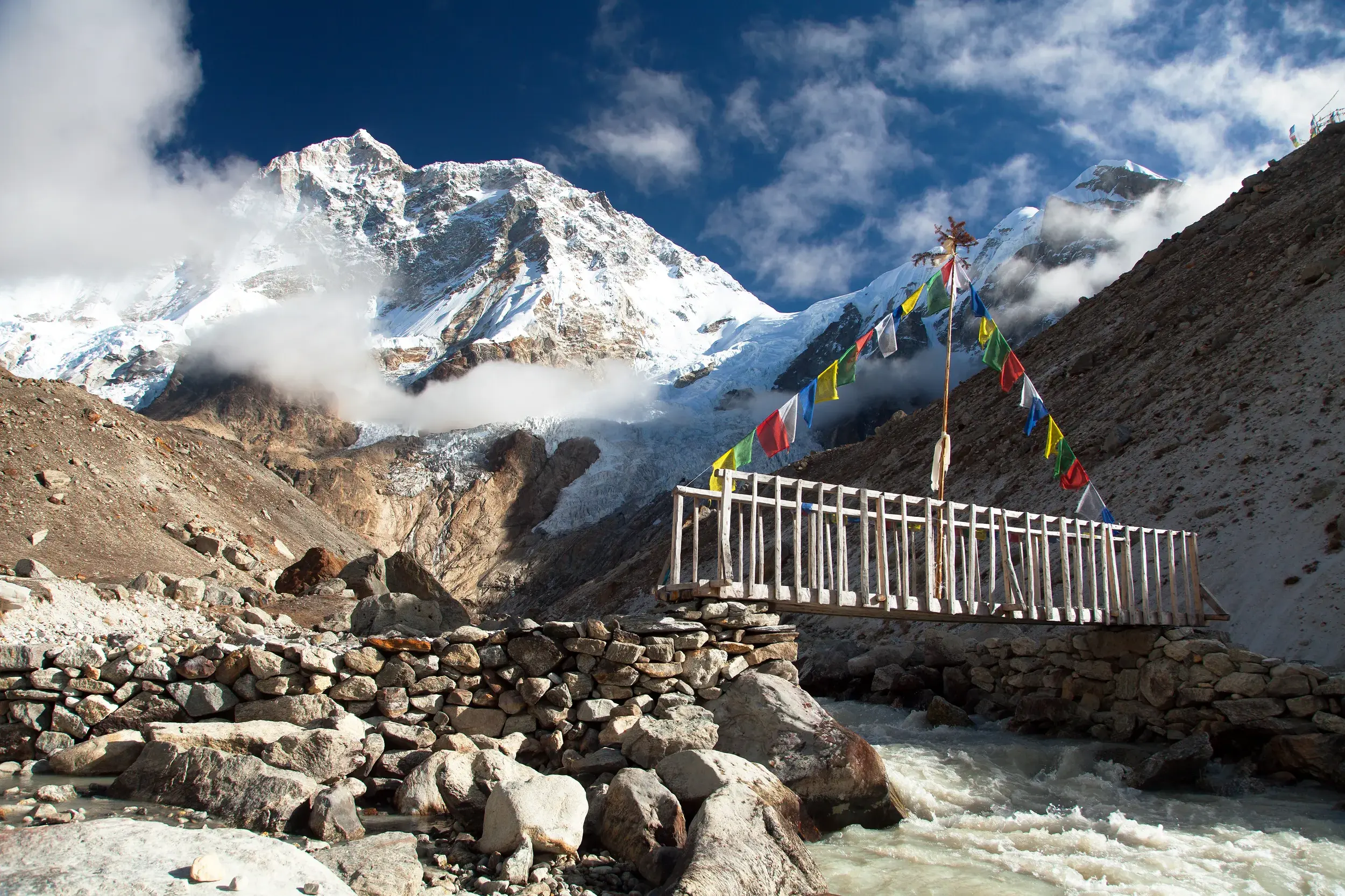 Eine Holzbrücke mit bunten Gebetsfahnen führt über einen Fluss beim Trekking zum Makalu-Basislager vor einem schneebedeckten Gipfel.