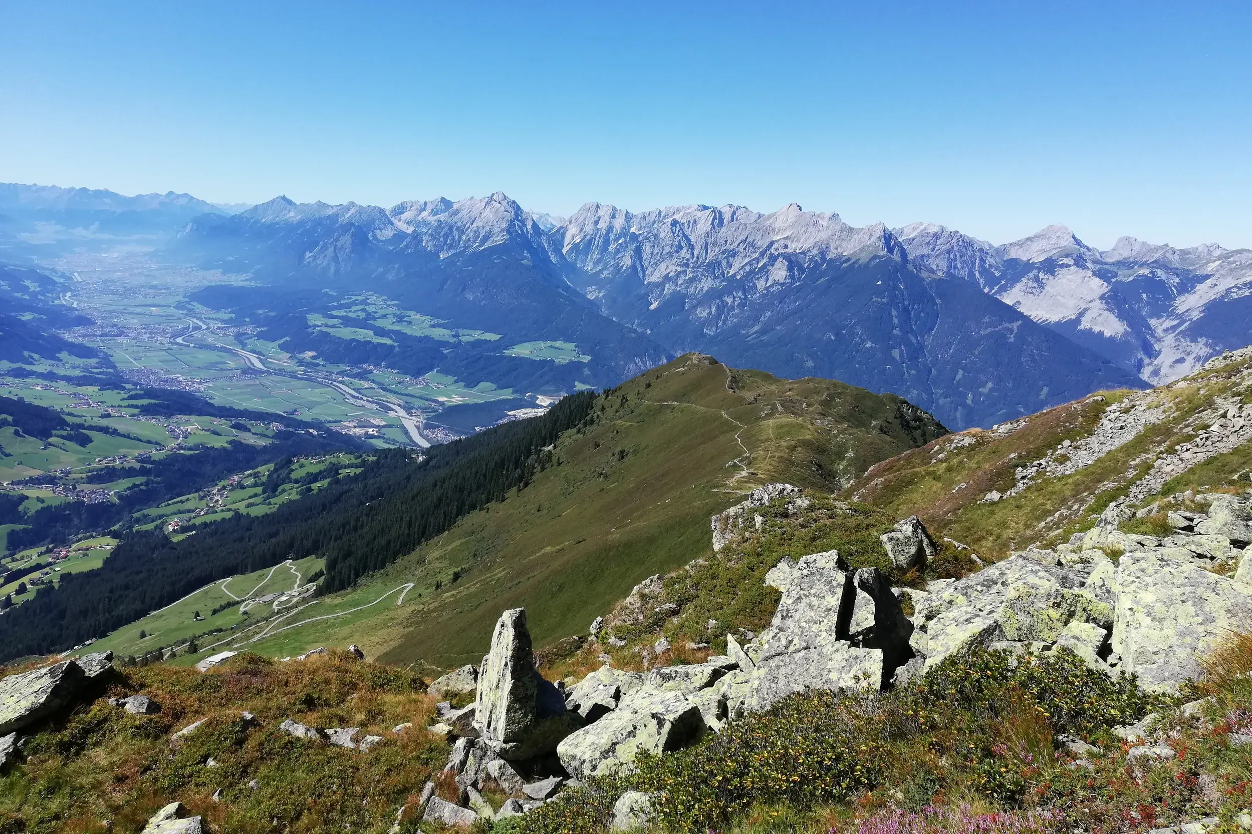 Sonnige Panoramasicht von einem Wanderweg über das weite Zillertal auf die schroffen Gipfel der Zillertaler Alpen.