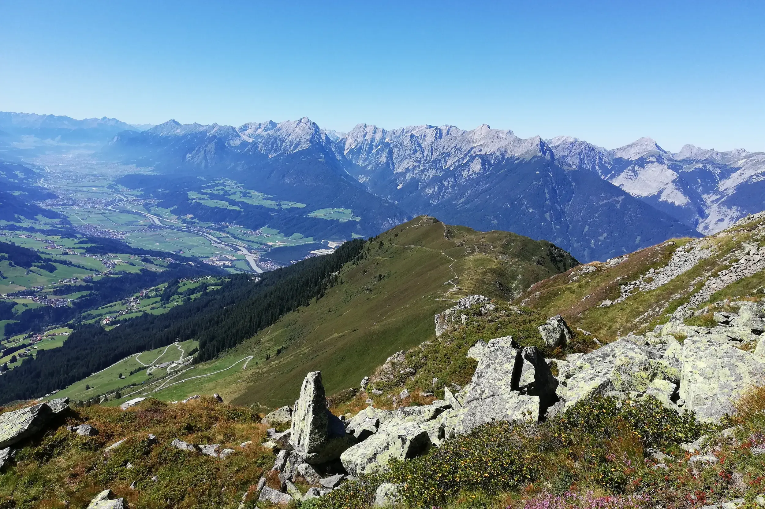 Sonnige Panoramasicht von einem Wanderweg über das weite Zillertal auf die schroffen Gipfel der Zillertaler Alpen.