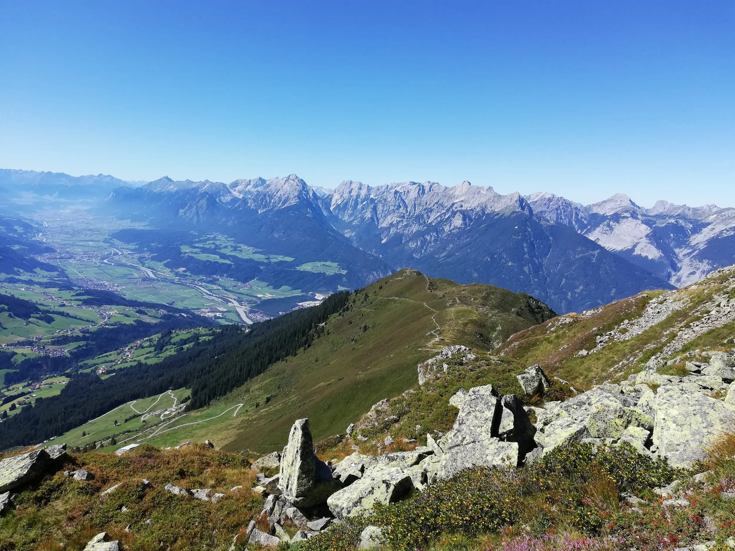 Sonnige Panoramasicht von einem Wanderweg über das weite Zillertal auf die schroffen Gipfel der Zillertaler Alpen.