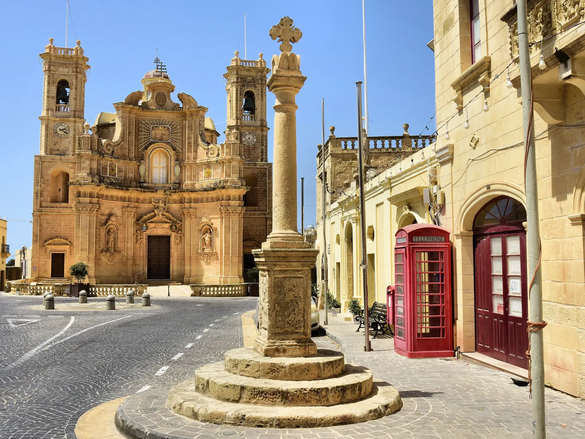 Prachtvolle Barockkirche aus Sandstein auf einem gepflasterten Platz auf Gozo, daneben eine typisch rote Telefonzelle.