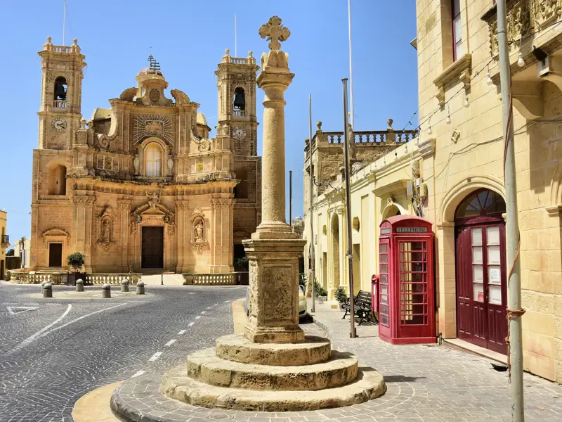 Prachtvolle Barockkirche aus Sandstein auf einem gepflasterten Platz auf Gozo, daneben eine typisch rote Telefonzelle.