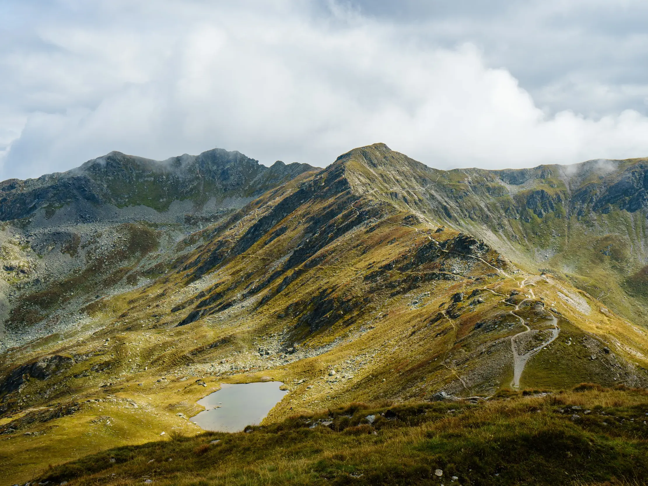 Ein Wanderpfad schlängelt sich über den grasbewachsenen Grat des Karnischen Höhenwegs, vorbei an einem Bergsee in den Alpen.