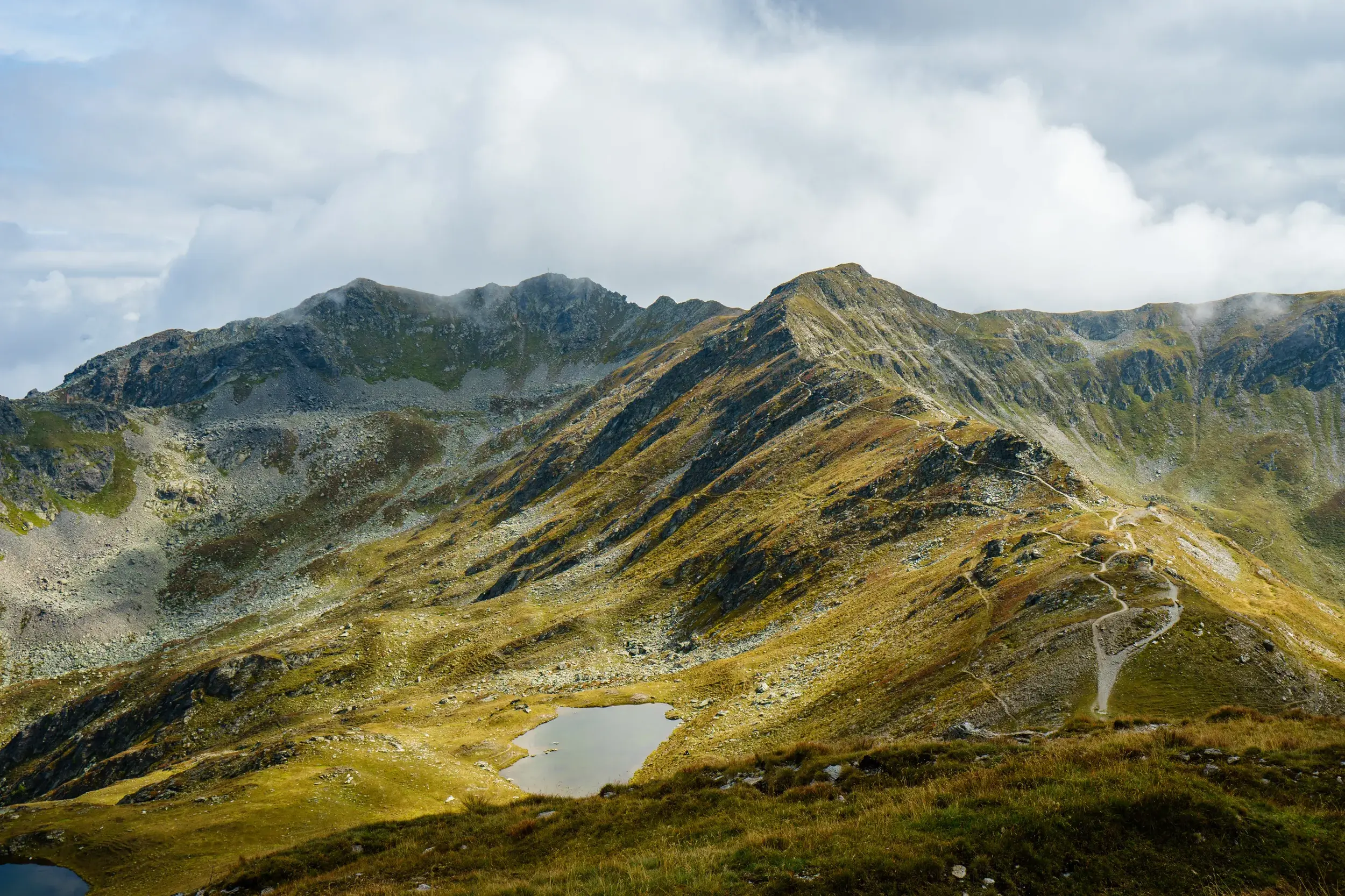 Ein Wanderpfad schlängelt sich über den grasbewachsenen Grat des Karnischen Höhenwegs, vorbei an einem Bergsee in den Alpen.