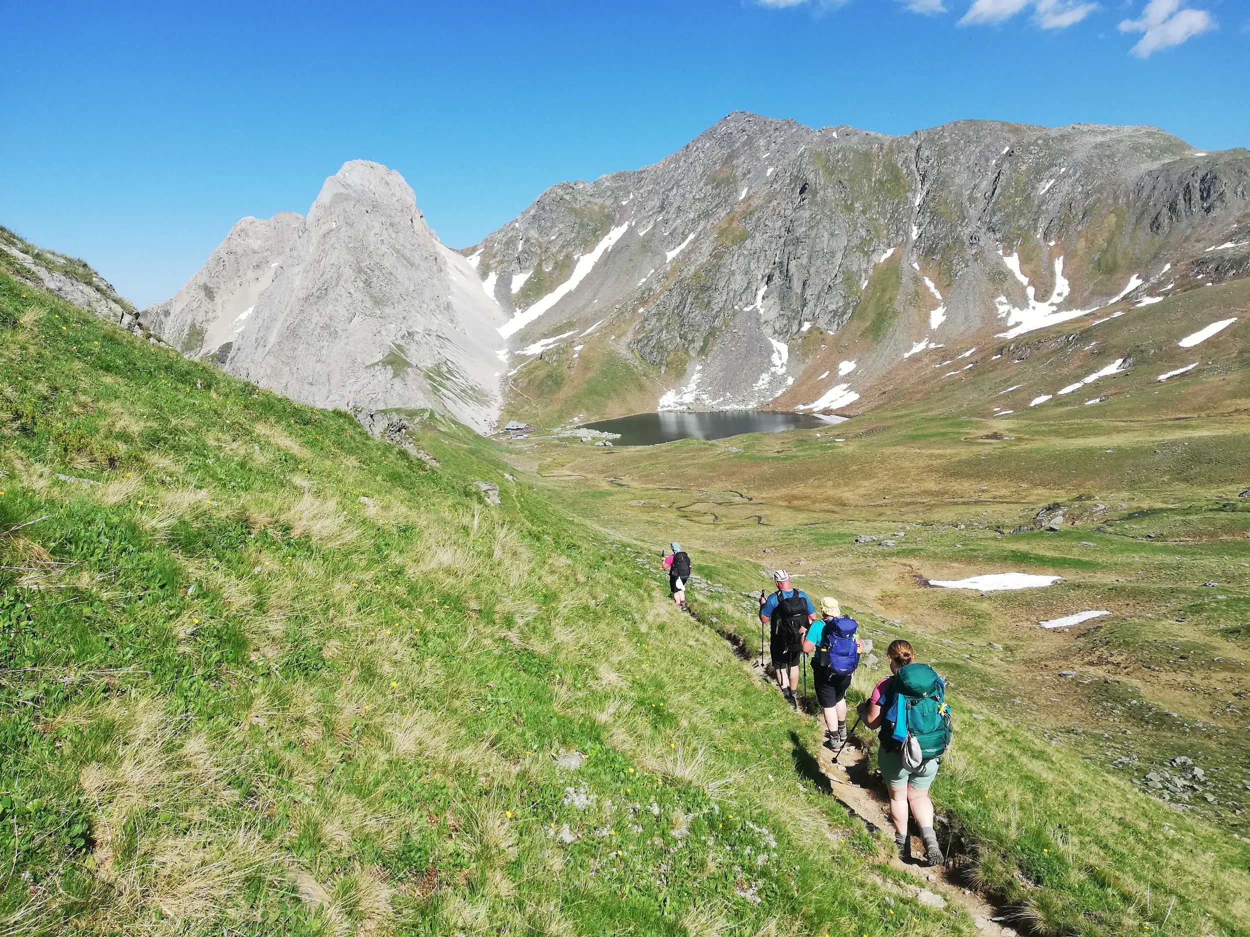 Eine Wandergruppe auf einem schmalen Pfad am Karnischen Höhenweg mit Blick auf einen Bergsee und die Gipfel der Karnischen Alpen.