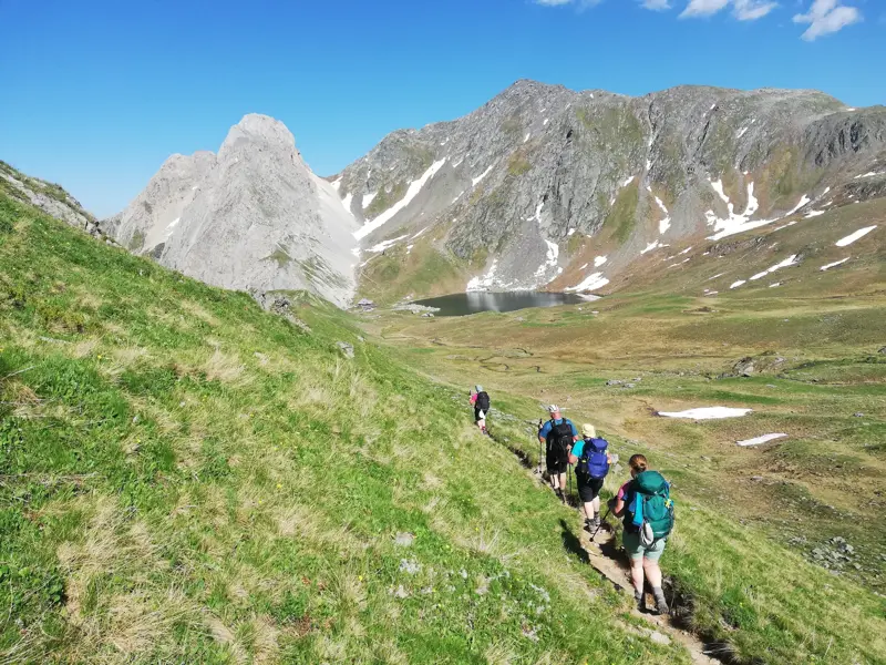 Kleine Wandergruppe beim Trekking auf einem schmalen Pfad am Karnischen Höhenweg mit Blick auf einen alpinen Bergsee im Tal.