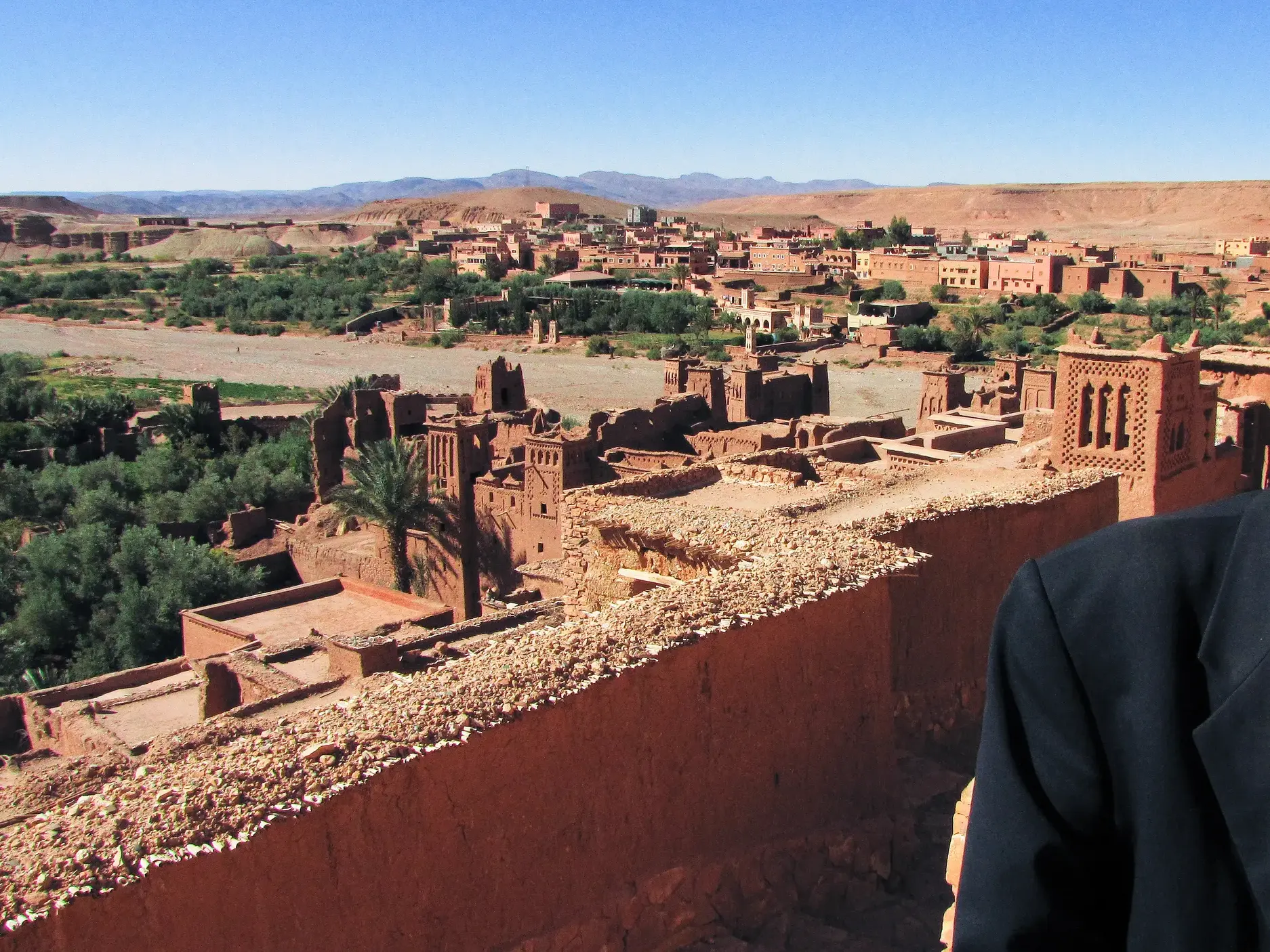 Ein lächelnder Berber mit Sonnenbrille vor dem sonnigen Panorama der UNESCO-geschützten Lehmburg Ait Benhaddou in Marokko.