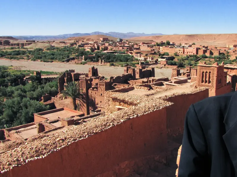 Ein lächelnder Berber mit Sonnenbrille vor dem sonnigen Panorama der UNESCO-geschützten Lehmburg Ait Benhaddou in Marokko.