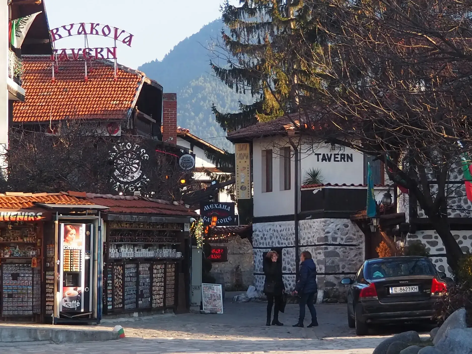 Eine Gasse in Bansko mit traditionellen Tavernen, einem Souvenirladen und Passanten vor der Bergkulisse des Pirin-Gebirges.
