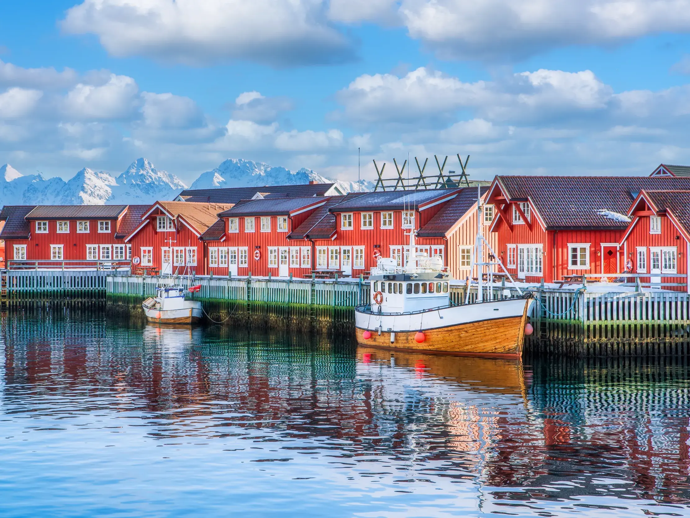 Rote Fischerhäuser und Boote im Hafen von Henningsvær auf den Lofoten spiegeln sich im Wasser vor schneebedeckten Bergen.