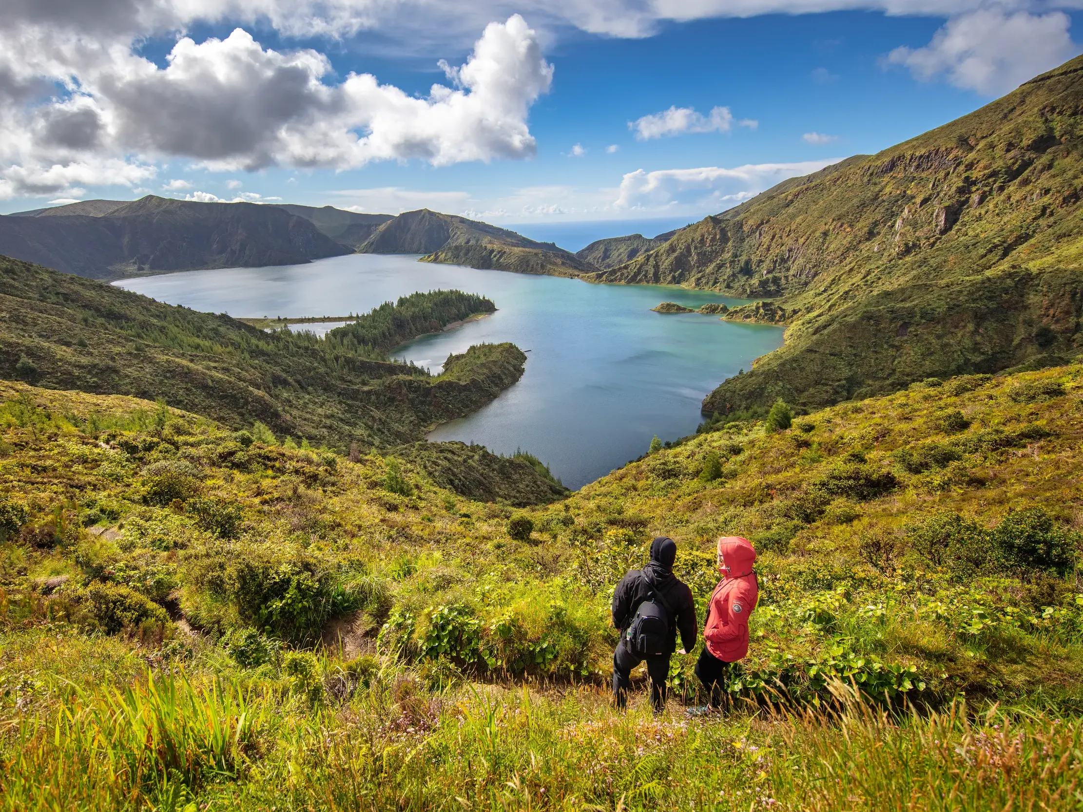 Zwei Wanderer blicken von einem grünen Hang auf den Vulkankratersee Lagoa do Fogo auf der Azoreninsel Sao Miguel.