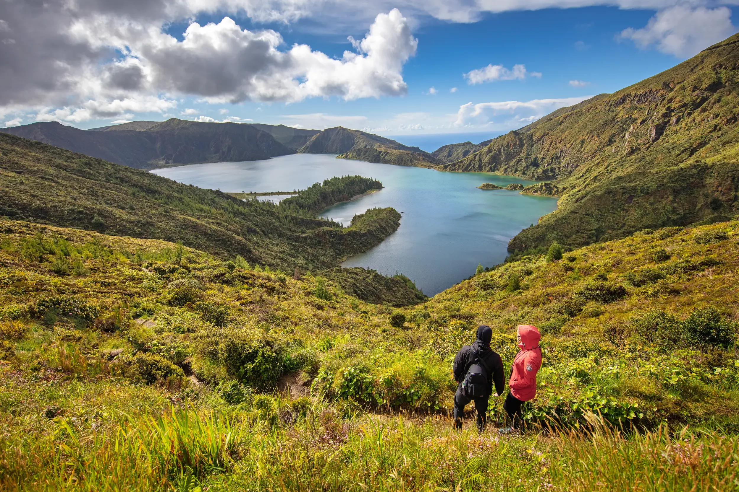 Zwei Wanderer blicken von einem grünen Hang auf den Vulkankratersee Lagoa do Fogo auf der Azoreninsel Sao Miguel.