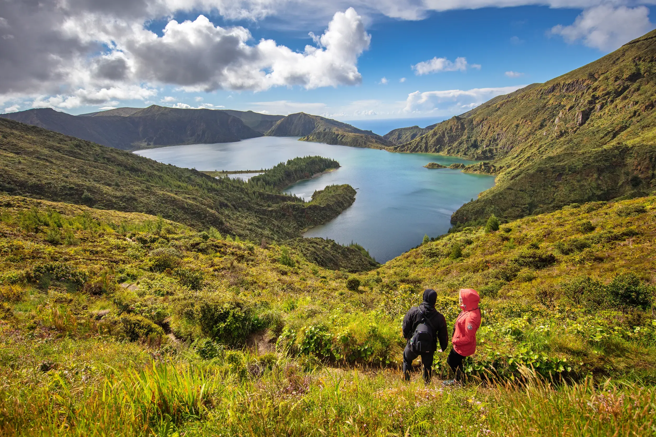 Zwei Wanderer blicken von einem grünen Hang auf den Vulkankratersee Lagoa do Fogo auf der Azoreninsel Sao Miguel.