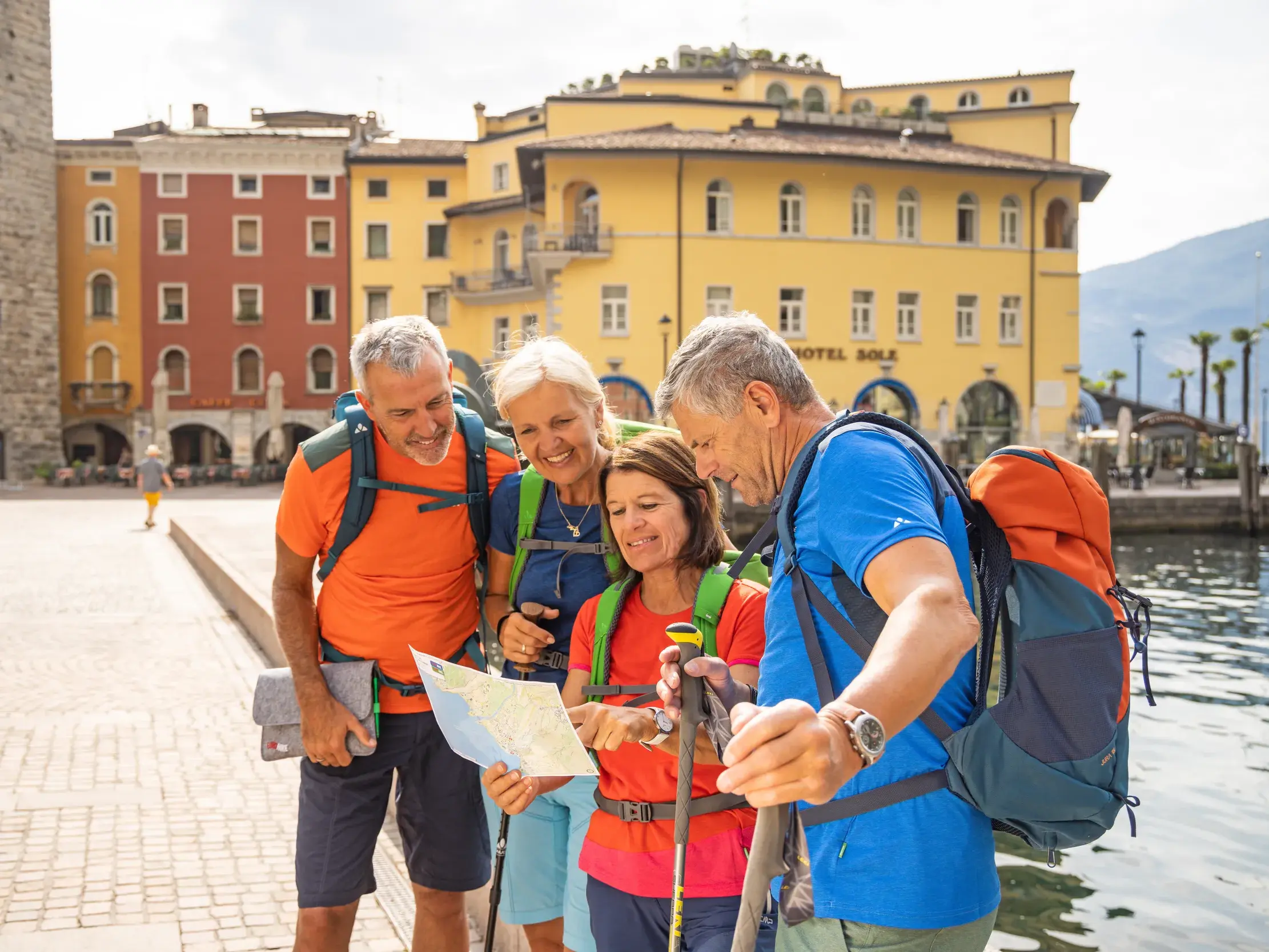 Vier aktive Wanderer in Wanderkleidung planen ihre Route auf einer Karte an der Uferpromenade von Riva del Garda am Gardasee.