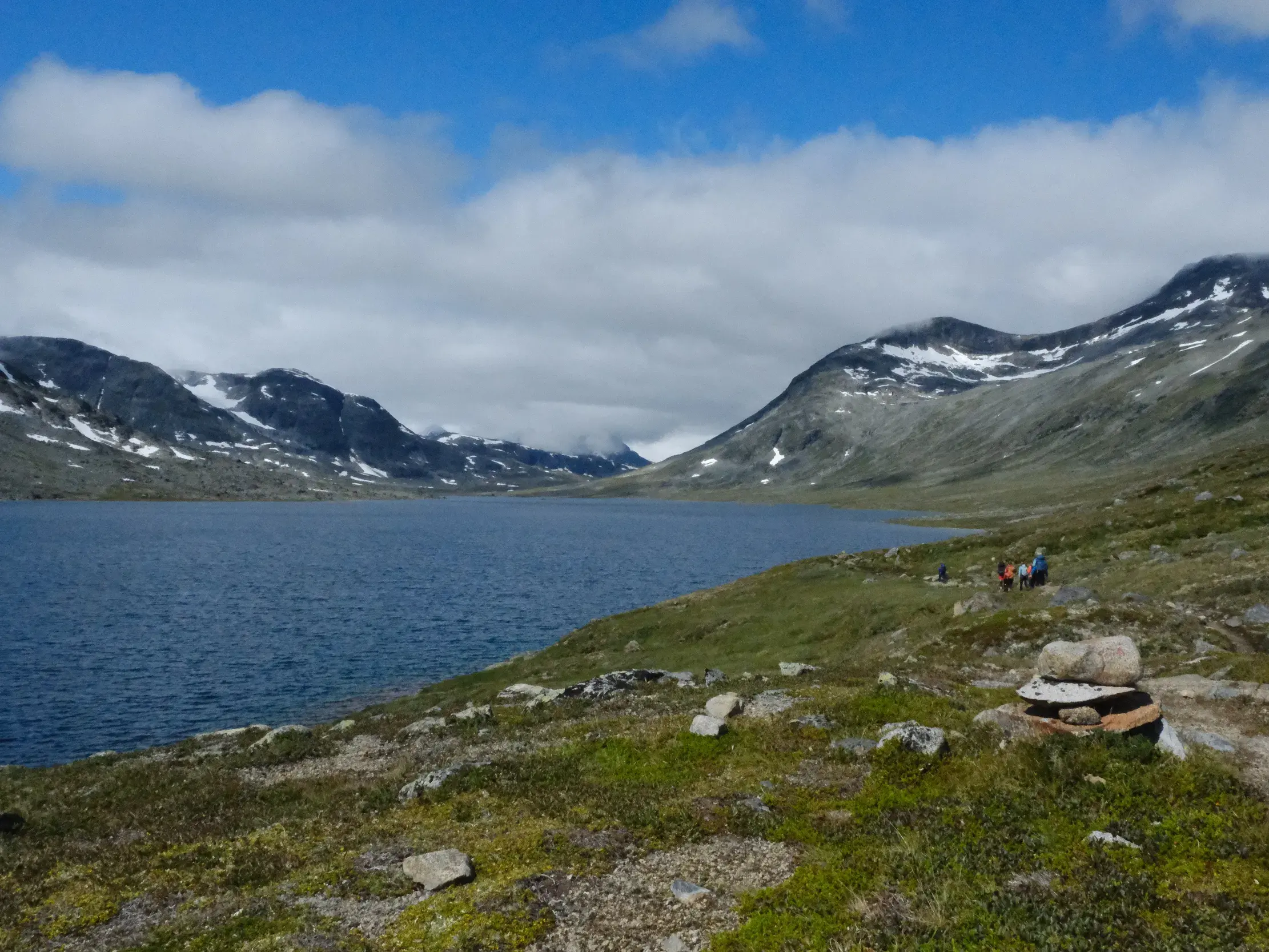 Eine kleine Wandergruppe auf einem Pfad am Ufer eines blauen Sees, umgeben von schneebedeckten Bergen im Jotunheimen Nationalpark.