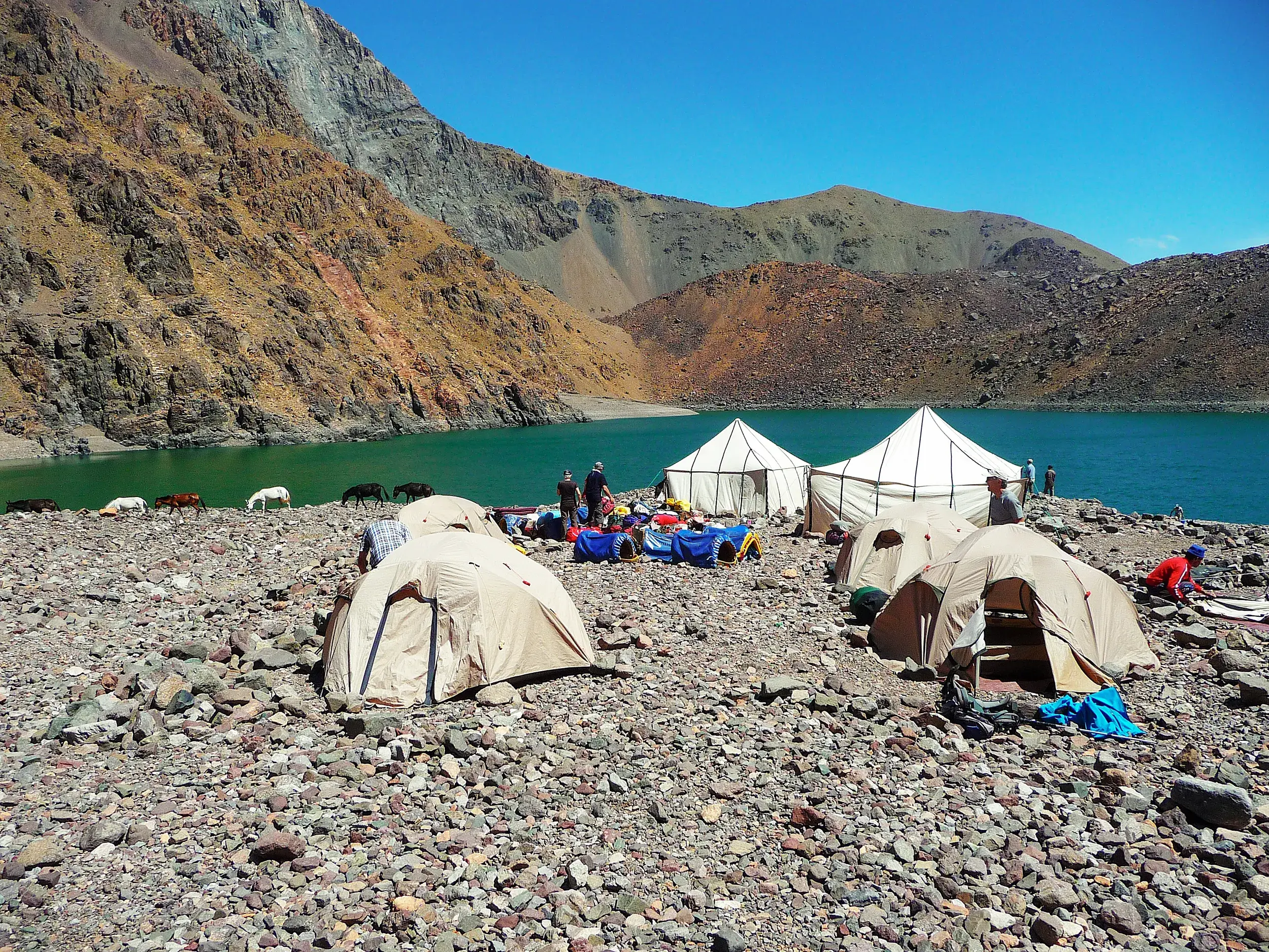Zelte einer Trekkinggruppe am felsigen Ufer des smaragdgrünen Lac d'Ifni, umgeben von den kargen Bergen des Hohen Atlas.