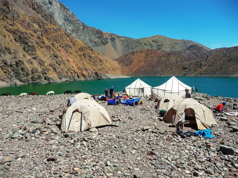 Zelte einer Trekkinggruppe am felsigen Ufer des smaragdgrünen Lac d'Ifni, umgeben von den kargen Bergen des Hohen Atlas.