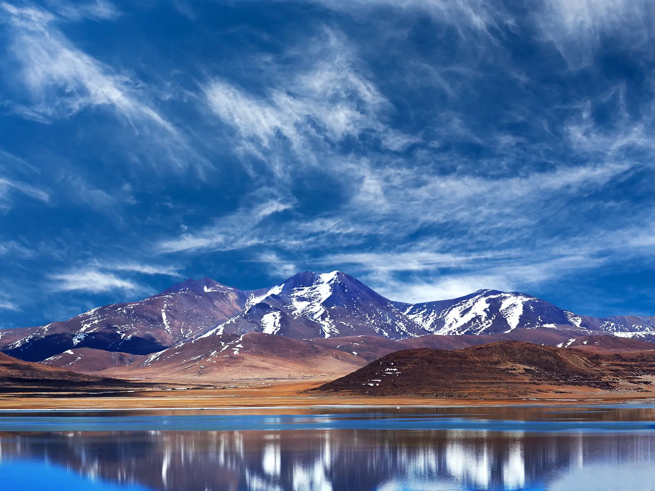 Schneebedeckte Gipfel des Himalaya spiegeln sich im tiefblauen Paiku-Tso See unter dramatischem Wolkenhimmel in Tibet.