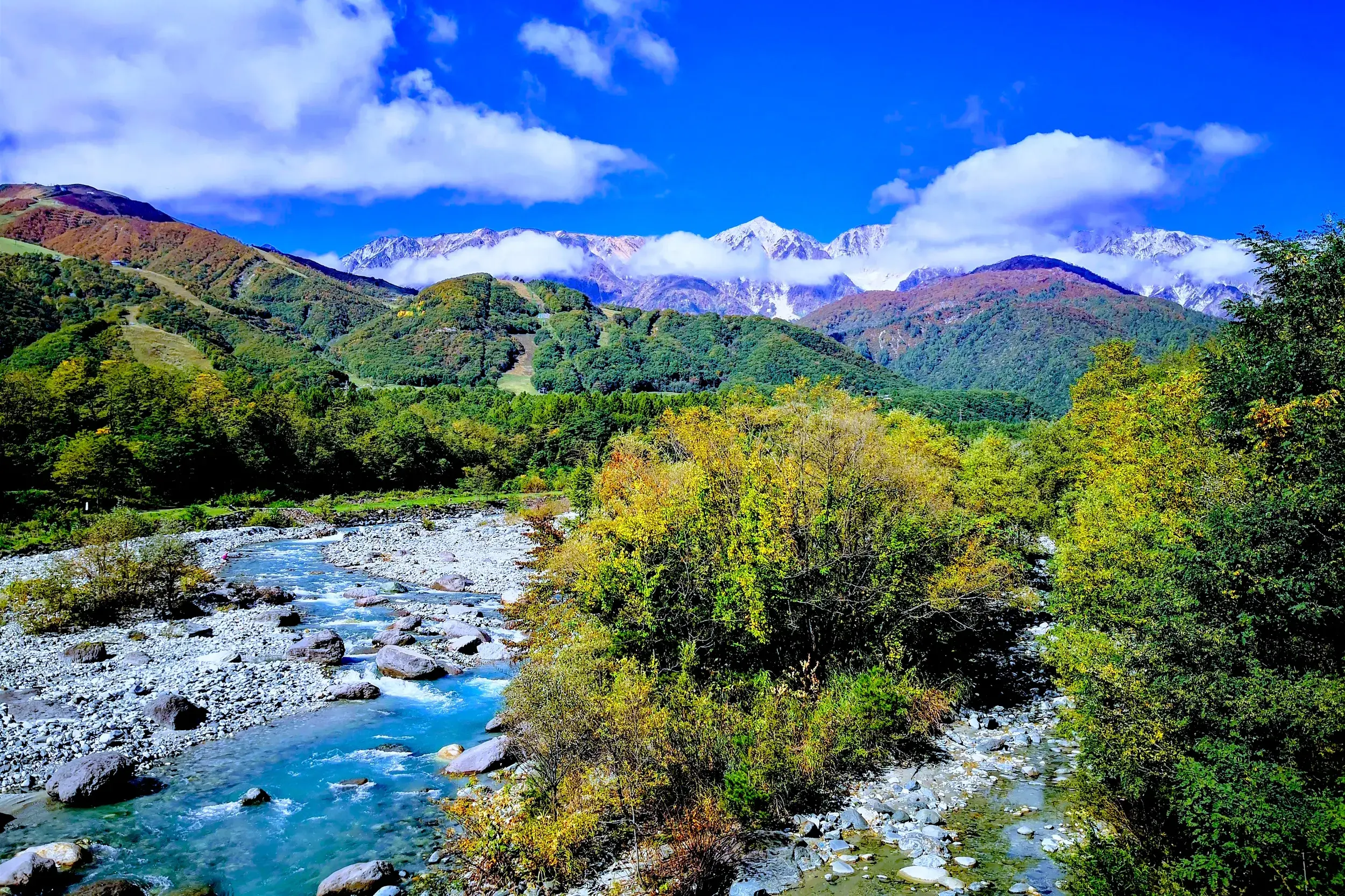 Türkisfarbener Gebirgsfluss im Herbstlaub vor der Kulisse der schneebedeckten Japanischen Alpen bei einer Wanderung in Hakuba.