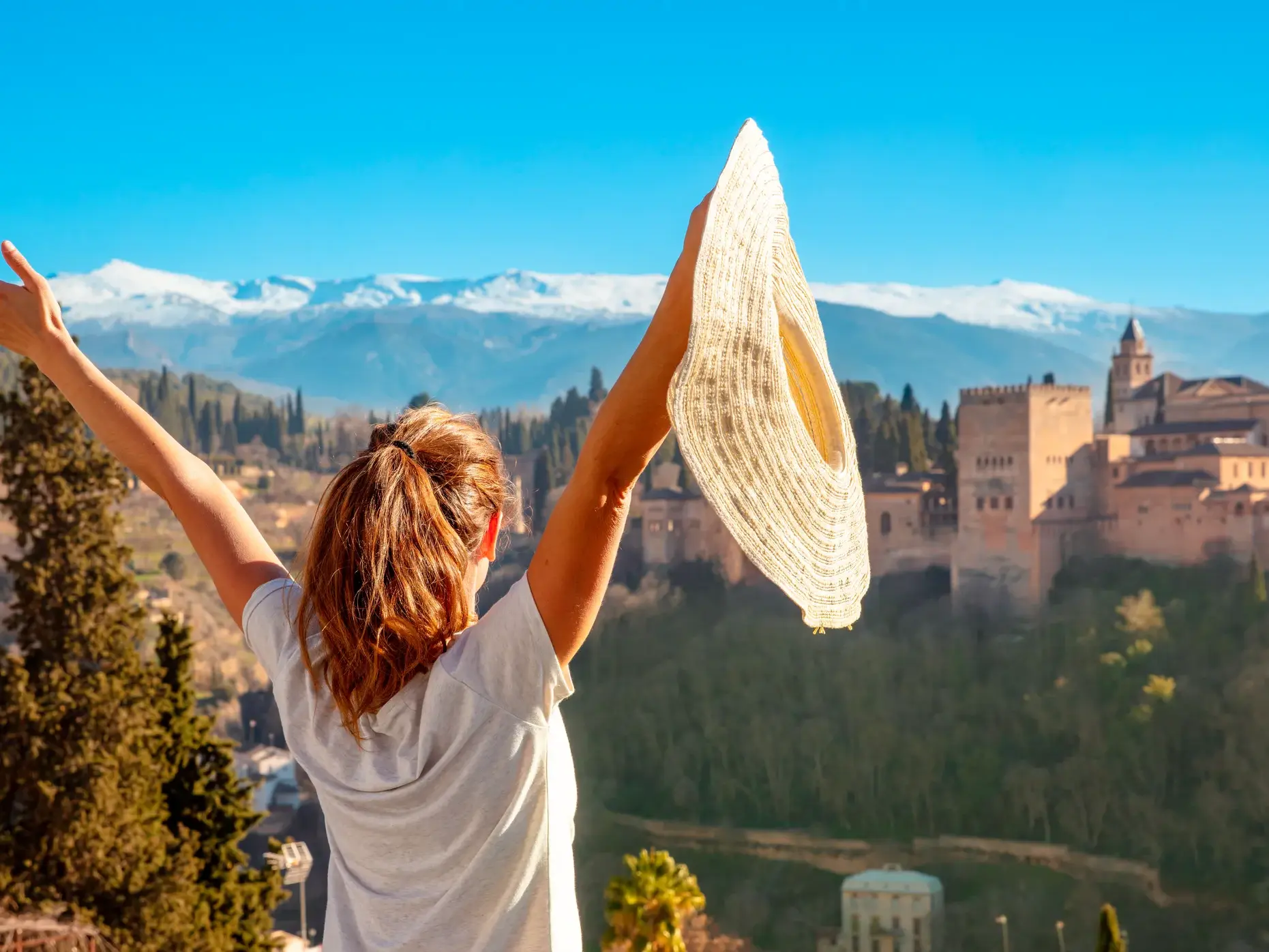 Eine junge Frau streckt jubelnd die Arme in die Luft und genießt den Ausblick auf die Alhambra vor der Sierra Nevada in Granada.
