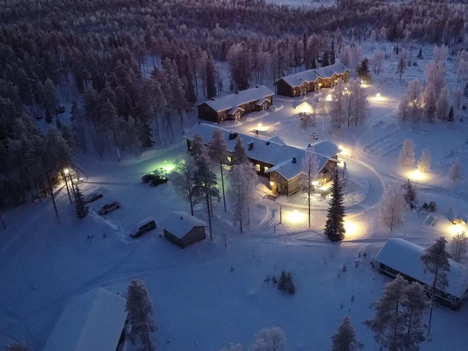 Die beleuchtete Saija Lodge aus der Luft, umgeben von tief verschneiten Wäldern in Finnisch Lappland bei Dämmerung.