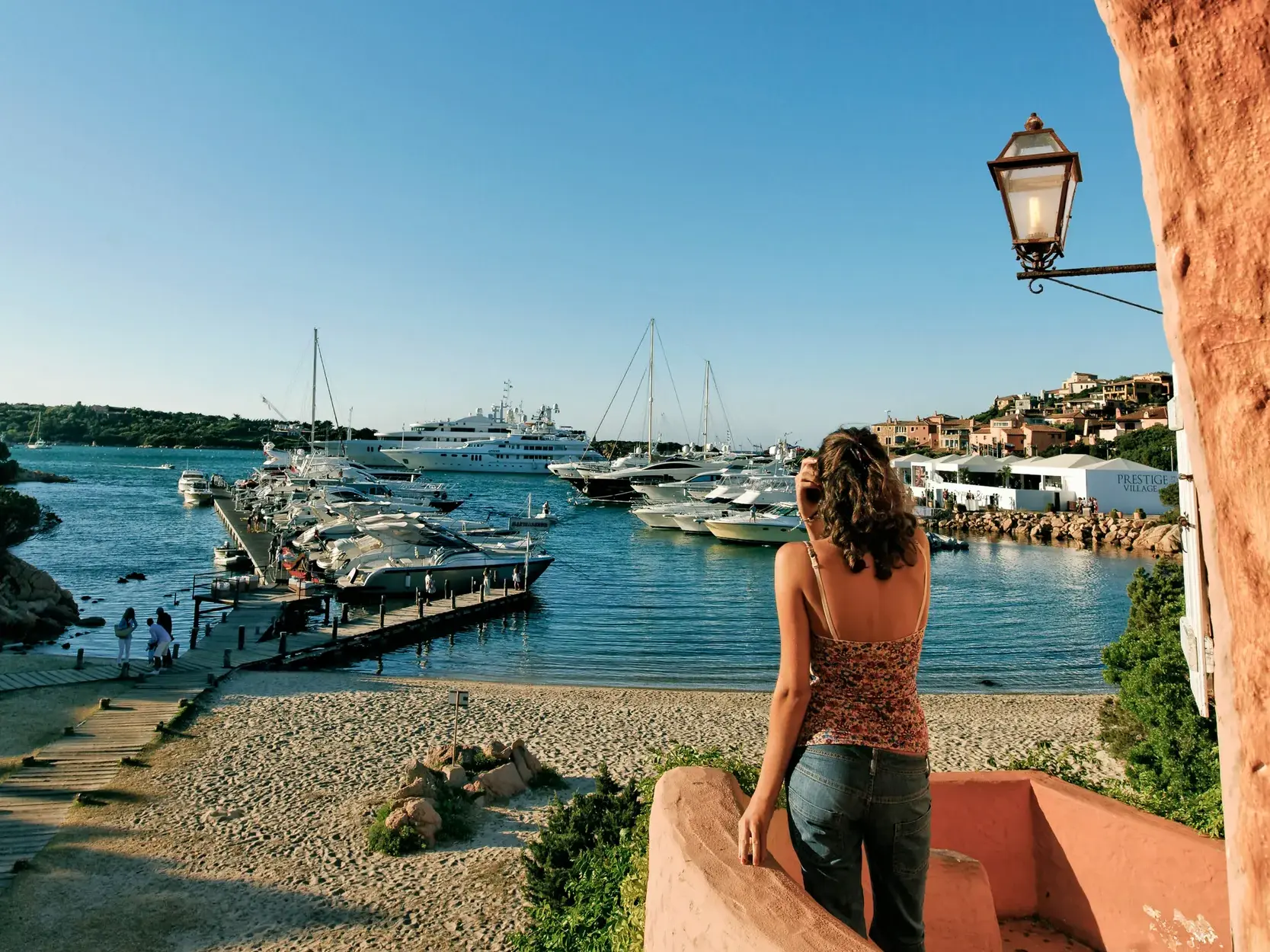 Eine junge Frau blickt von einem Balkon auf den luxuriösen Yachthafen von Porto Cervo an der Costa Smeralda in Sardinien.