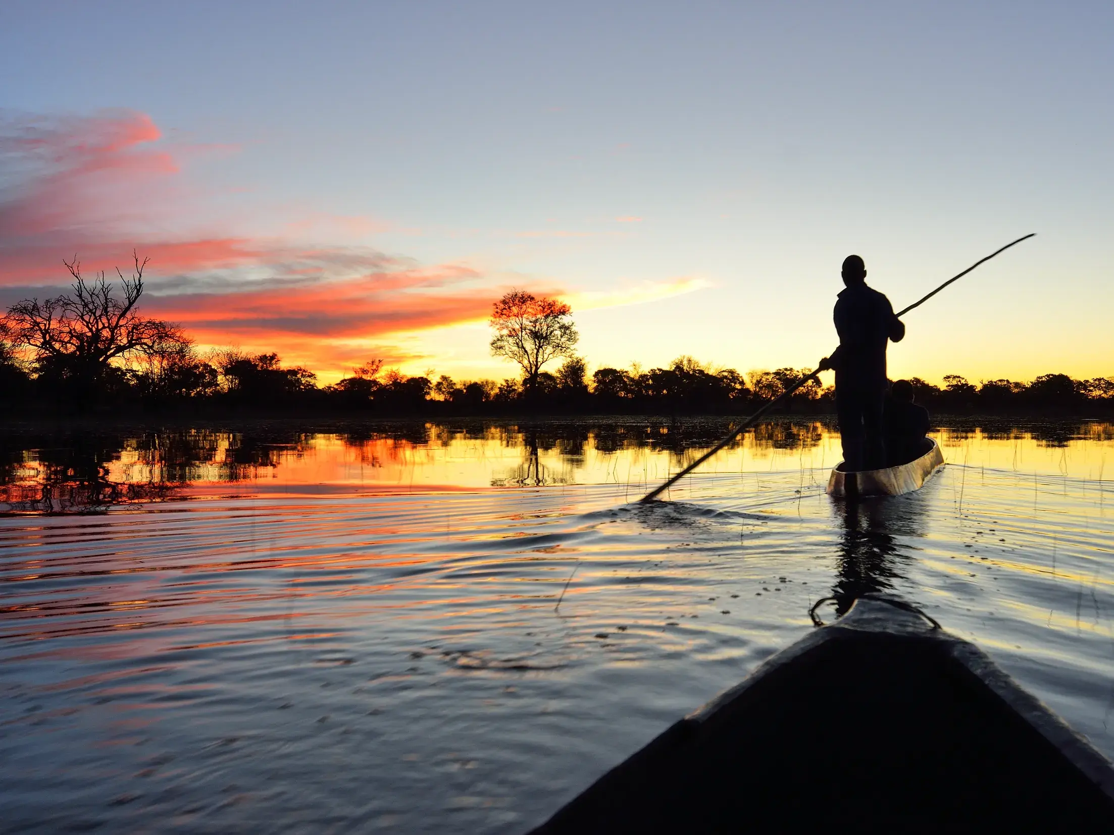 Ein Guide stakt einen Mokoro-Einbaum durch die Wasserwege des Okavangodeltas bei malerischem Sonnenuntergang in Botswana.
