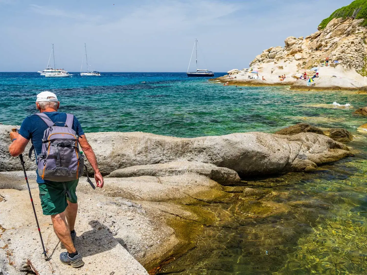Ein Wanderer mit Rucksack und Stöcken beim Trekking auf einem Felsenpfad entlang der türkisblauen Küste von Elba.