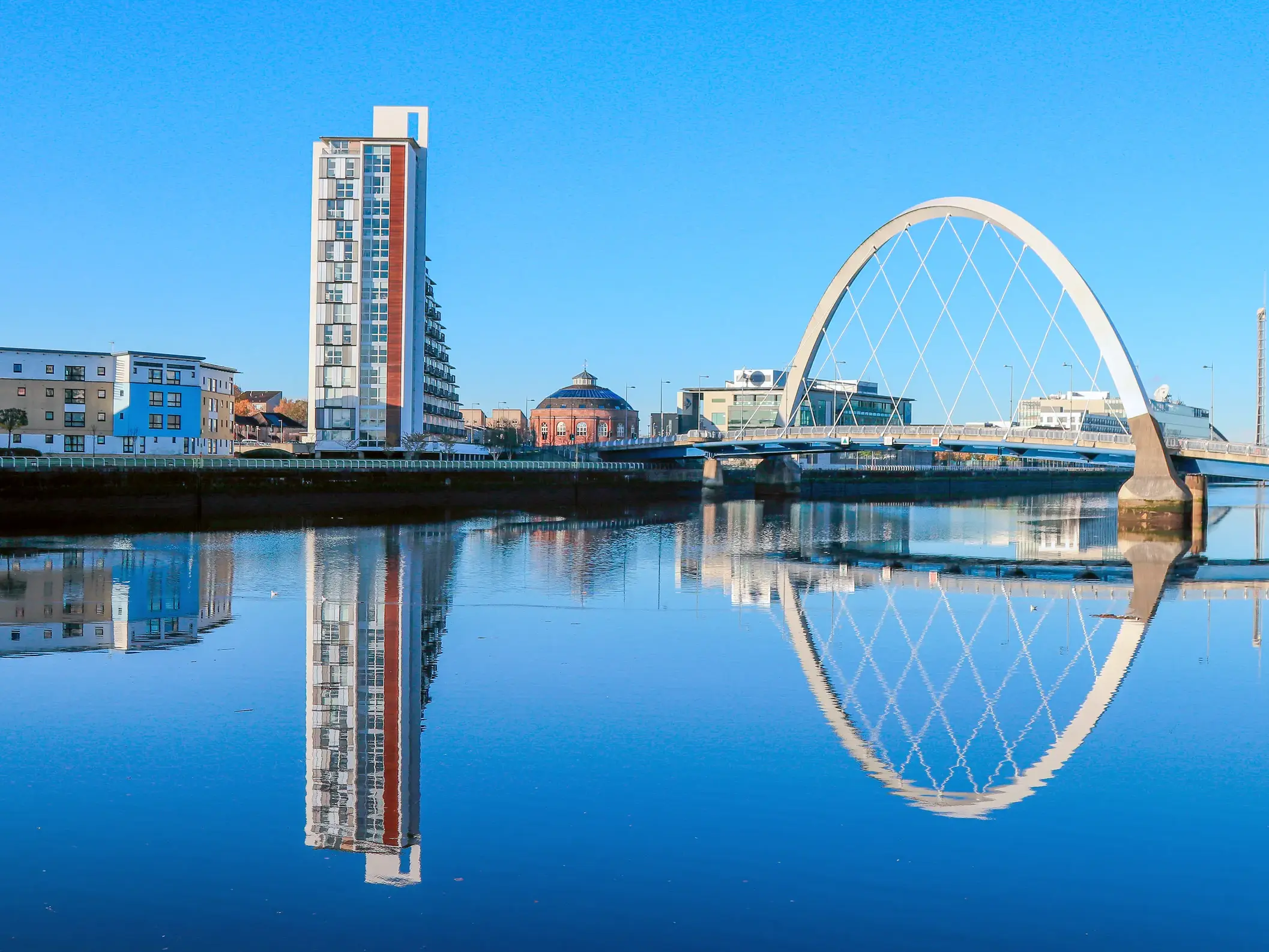 Die moderne Bogenbrücke Clyde Arc spiegelt sich im Fluss Clyde vor der Skyline von Glasgow bei blauem Himmel.