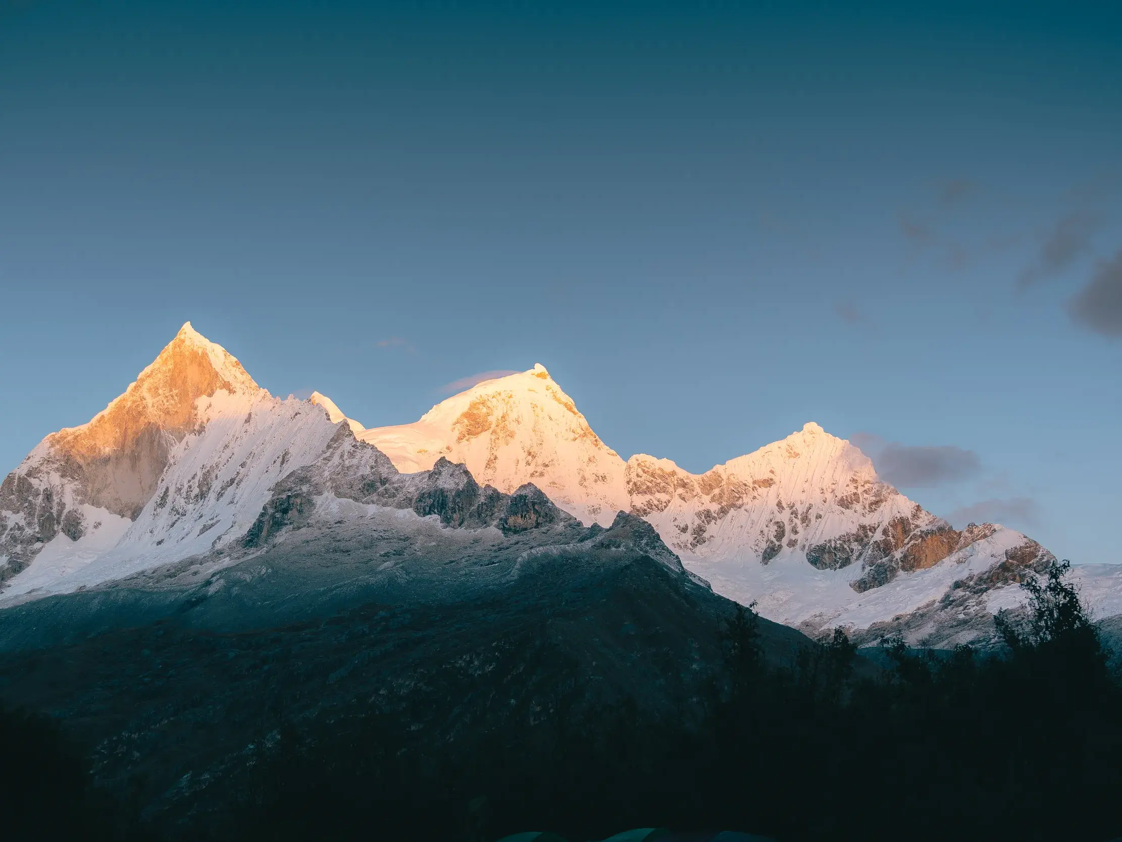 Schneebedeckte Gipfel der Cordillera Blanca in Peru leuchten im goldenen Licht des Sonnenaufgangs über einem dunklen Tal.
