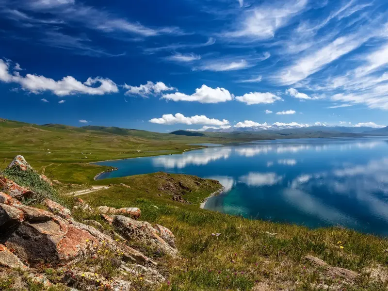 Panoramablick auf den spiegelglatten Songköl-See in Kirgistan, umgeben von grünen Weiden mit Jurten und den Bergen am Horizont.