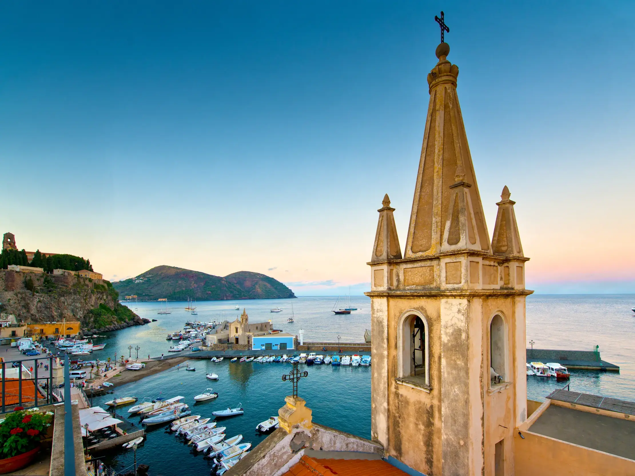 Blick von Lipari-Stadt über den Hafen Marina Corta mit ankernden Booten zum Kastell und der Insel Vulcano in der Abenddämmerung.