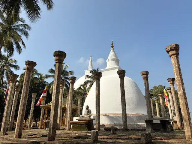 Antike Steinsäulen umrahmen eine weiße Stupa und Buddha-Statue in der heiligen Tempelanlage von Anuradhapura in Sri Lanka.