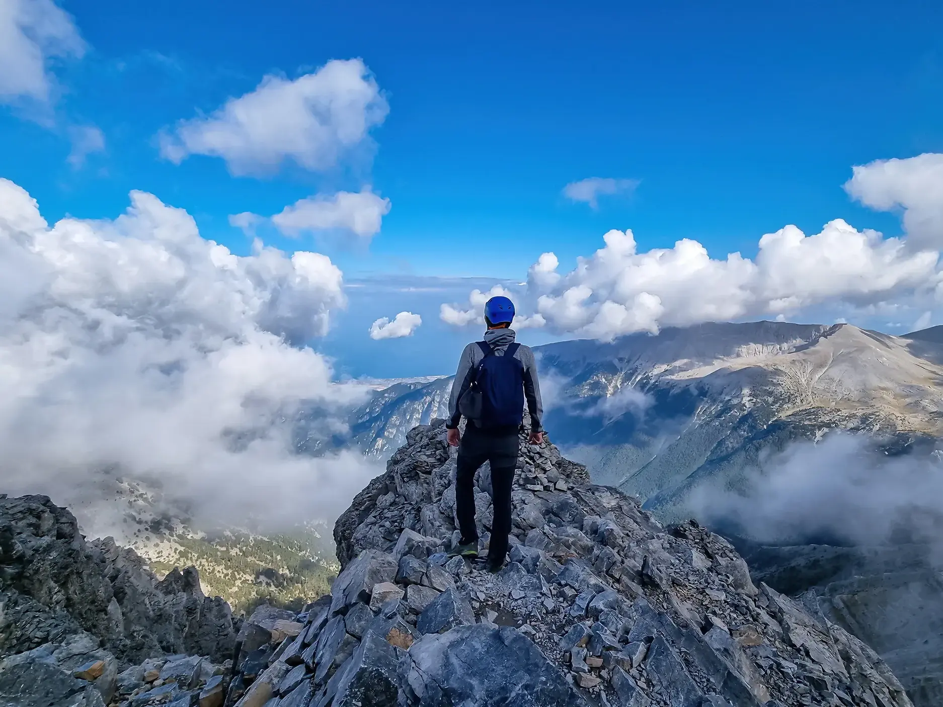 Ein Trekker genießt nach der Gipfelbesteigung die Aussicht vom felsigen Grat des Olymp-Massivs über die Wolken.