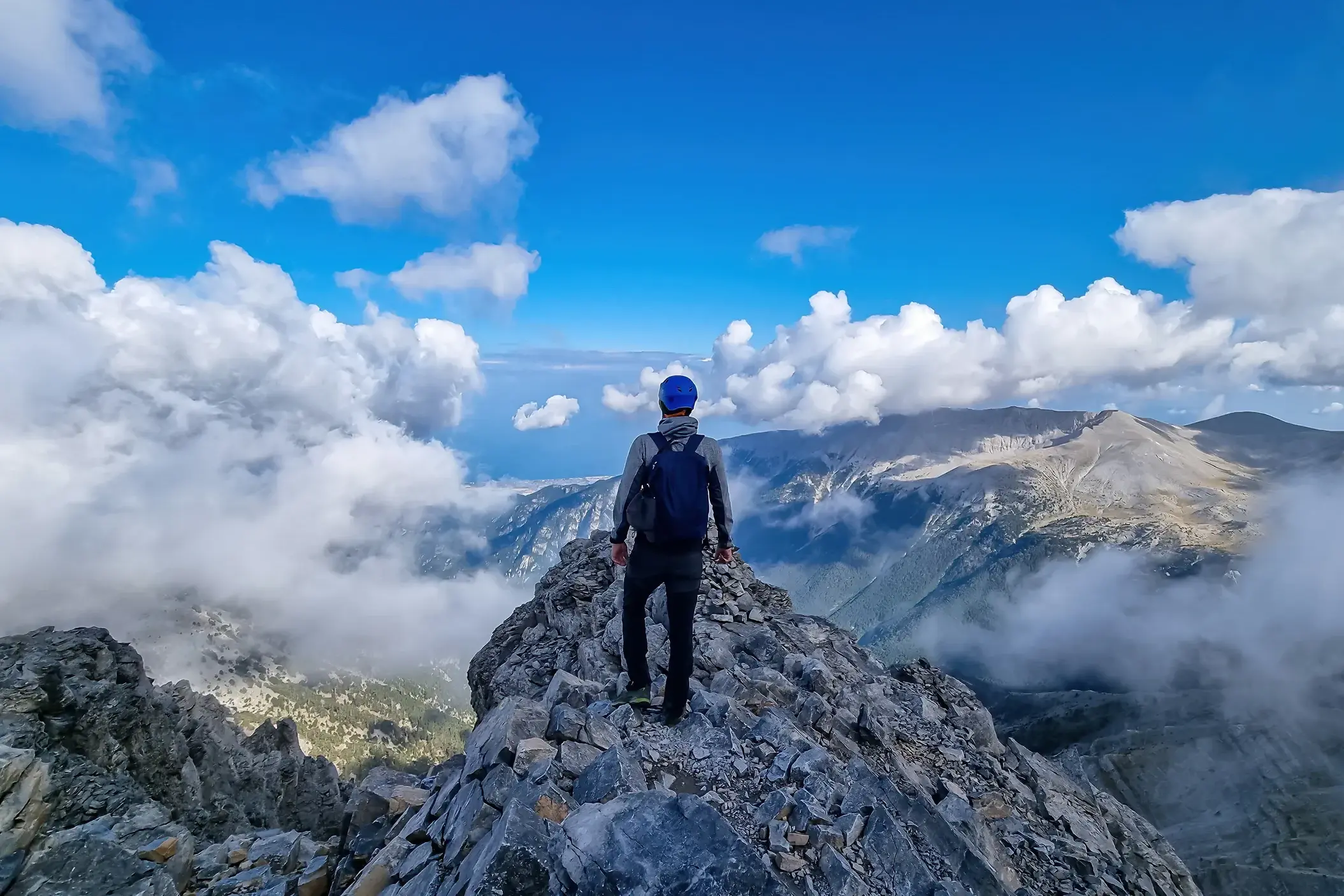 Ein Trekker genießt nach der Gipfelbesteigung die Aussicht vom felsigen Grat des Olymp-Massivs über die Wolken.