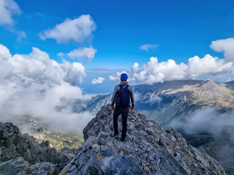 Ein Trekker genießt nach der Gipfelbesteigung die Aussicht vom felsigen Grat des Olymp-Massivs über die Wolken.