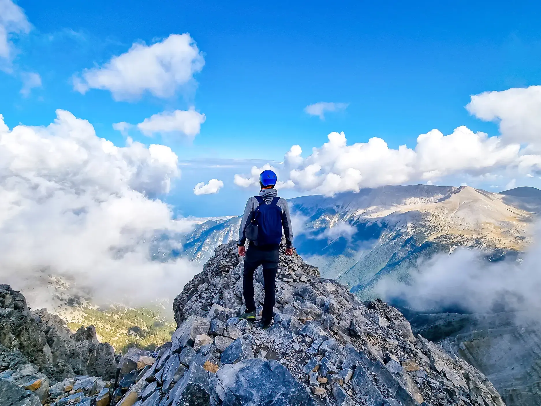 Wanderer mit Rucksack auf einem schmalen Felsgrat im Olymp-Massiv mit Blick über die Wolken auf das Ägäische Meer in Griechenland.