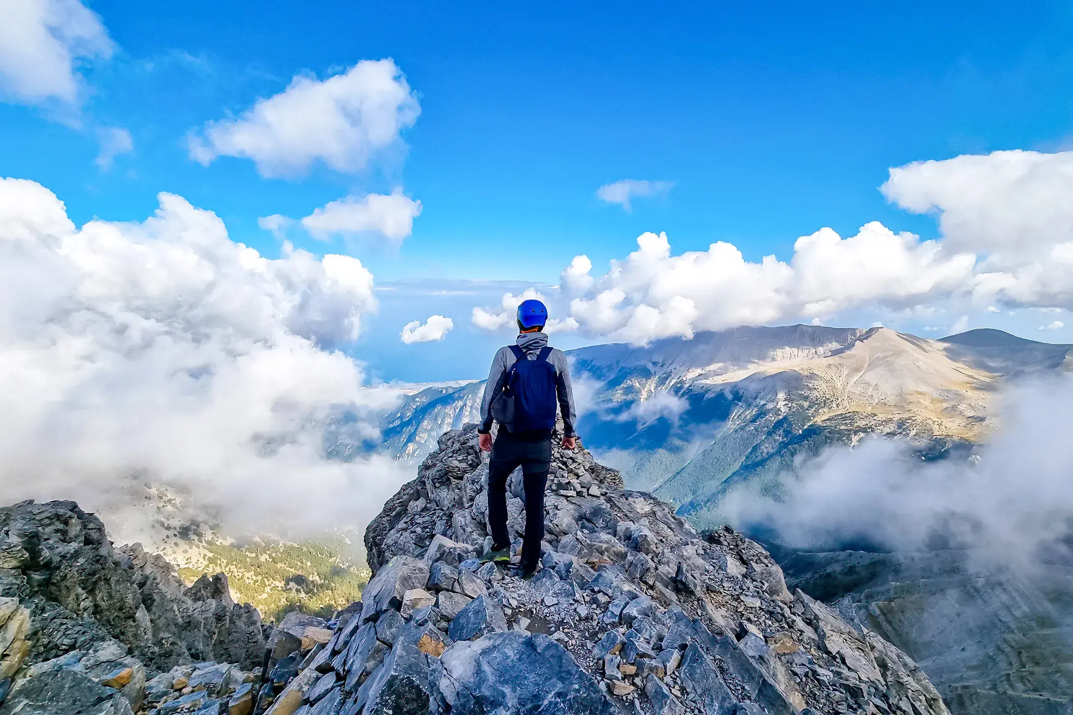 Wanderer mit Rucksack auf einem schmalen Felsgrat im Olymp-Massiv mit Blick über die Wolken auf das Ägäische Meer in Griechenland.