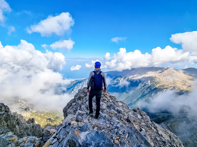 Wanderer mit Rucksack auf einem schmalen Felsgrat im Olymp-Massiv mit Blick über die Wolken auf das Ägäische Meer in Griechenland.
