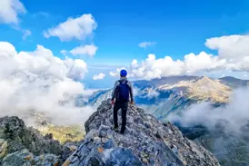 Wanderer mit Rucksack auf einem schmalen Felsgrat im Olymp-Massiv mit Blick über die Wolken auf das Ägäische Meer in Griechenland.