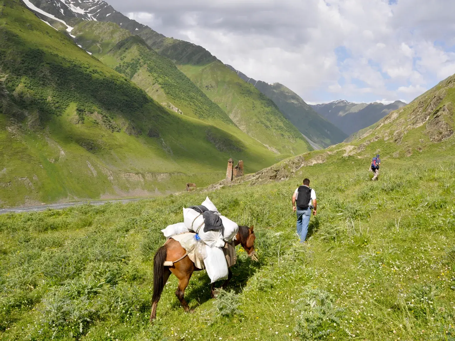 Trekking in Tuschetien: Wanderer und Packpferd in einem grünen Tal mit alten Wehrtürmen im Kaukasus.