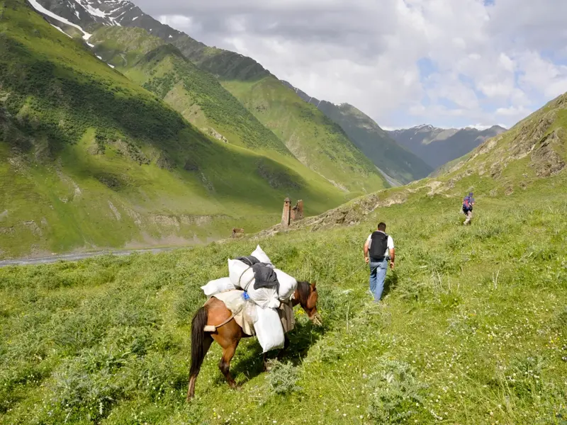 Trekking in Tuschetien: Wanderer und Packpferd in einem grünen Tal mit alten Wehrtürmen im Kaukasus.