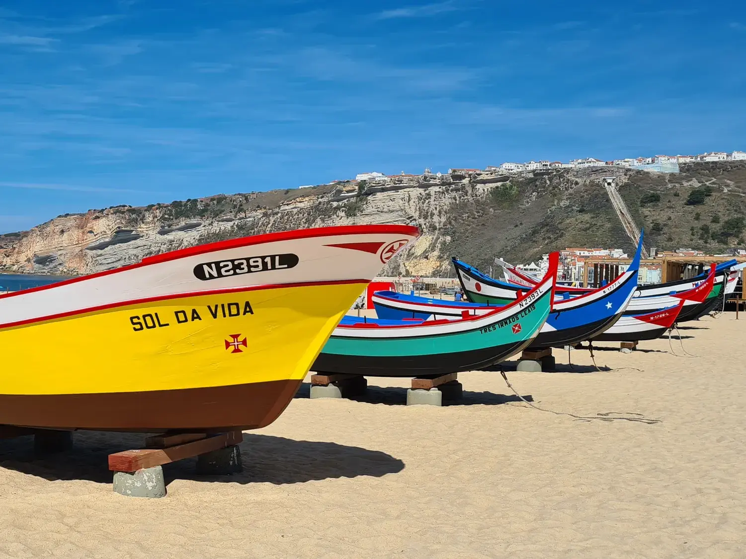 Farbenfrohe, traditionelle Fischerboote stehen am Sandstrand von Nazaré vor der Kulisse der Steilküste Portugals.