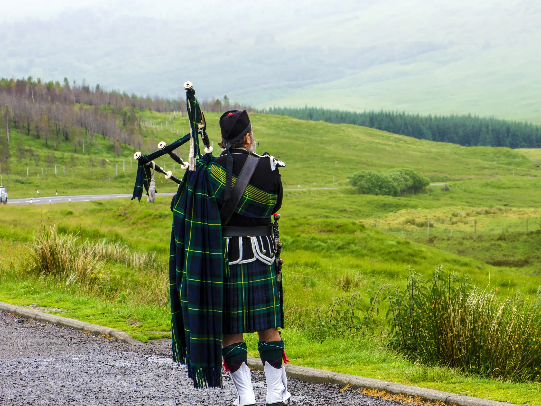 Ein Mann in schottischer Tracht mit Dudelsack blickt über die nebligen, grünen Weiten der schottischen Highlands.