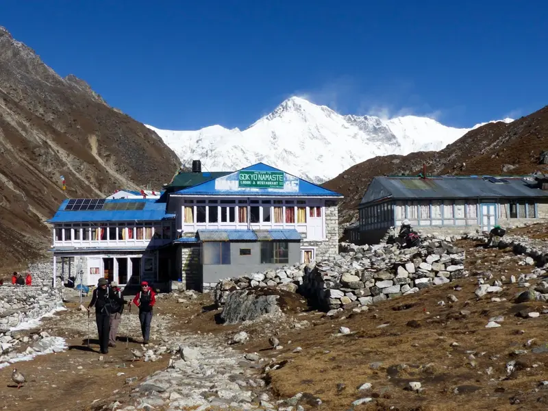 Trekker wandern an der Gokyo Lodge vorbei mit Panoramablick auf den schneebedeckten Achttausender Cho Oyu in Nepal.
