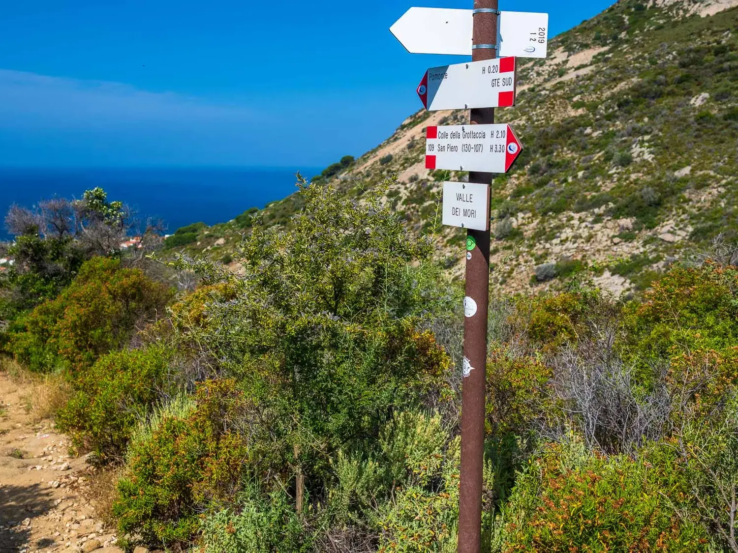 Ein roter Wanderwegweiser auf dem GTE auf Elba, umgeben von grüner Macchia, mit Panoramablick auf das Mittelmeer.