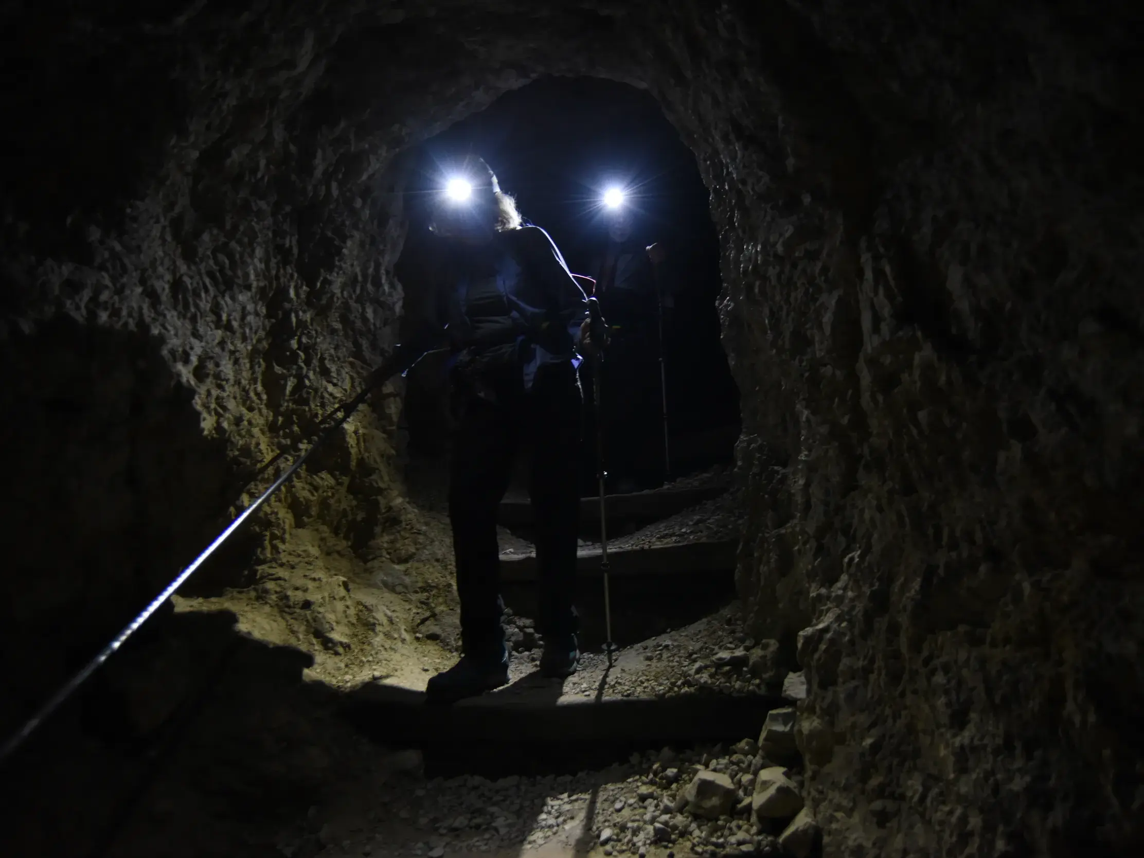 Wanderer steigen mit Stirnlampen durch den historischen Felstunnel vom Lagazuoi hinab zum Passo Falzarego in den Dolomiten.