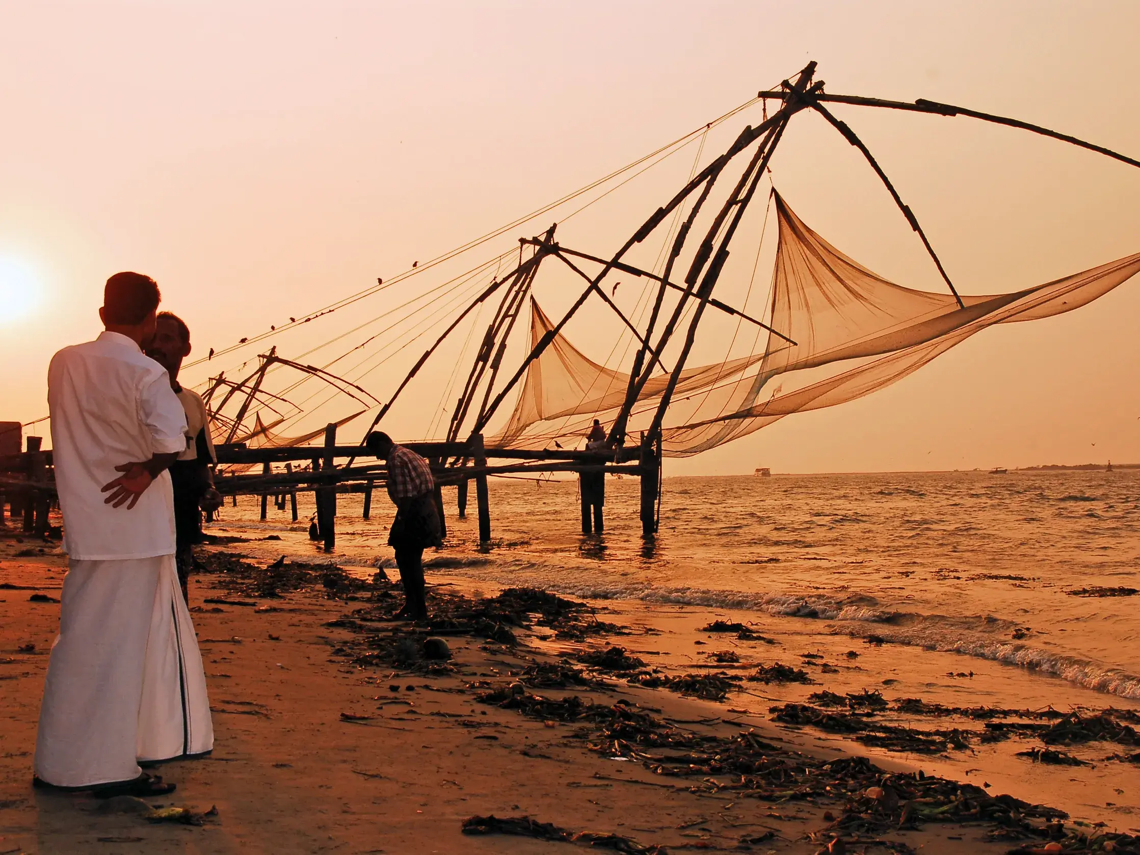 Männer in weißer Kleidung betrachten die großen Chinesischen Fischernetze am Strand von Kochi bei malerischem Sonnenuntergang.